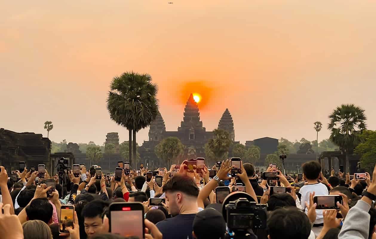 Angkor Wat temple at sunset with tourists capturing the view on their smartphones.