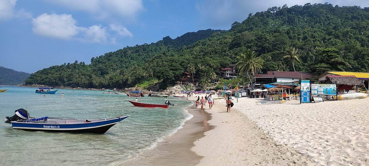 Scenic tropical beach with boats, white sand, and lush green hills in the background.